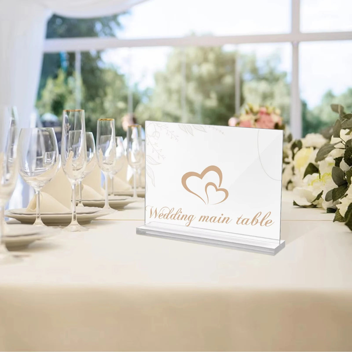A clear acrylic double-sided sign holder displaying the text Wedding main table on a white tablecloth with glassware in the b