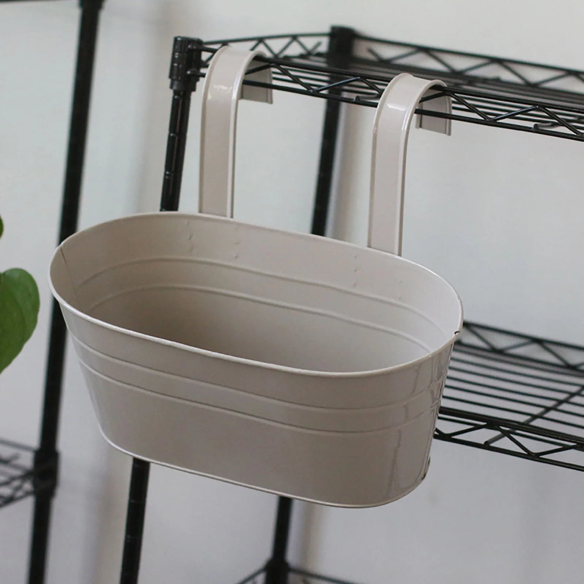 A modern boho metal hanging planter in off-white is displayed on a black wire shelf.