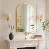 An arched wall mirror with an aluminum frame hangs above a marble fireplace mantel.