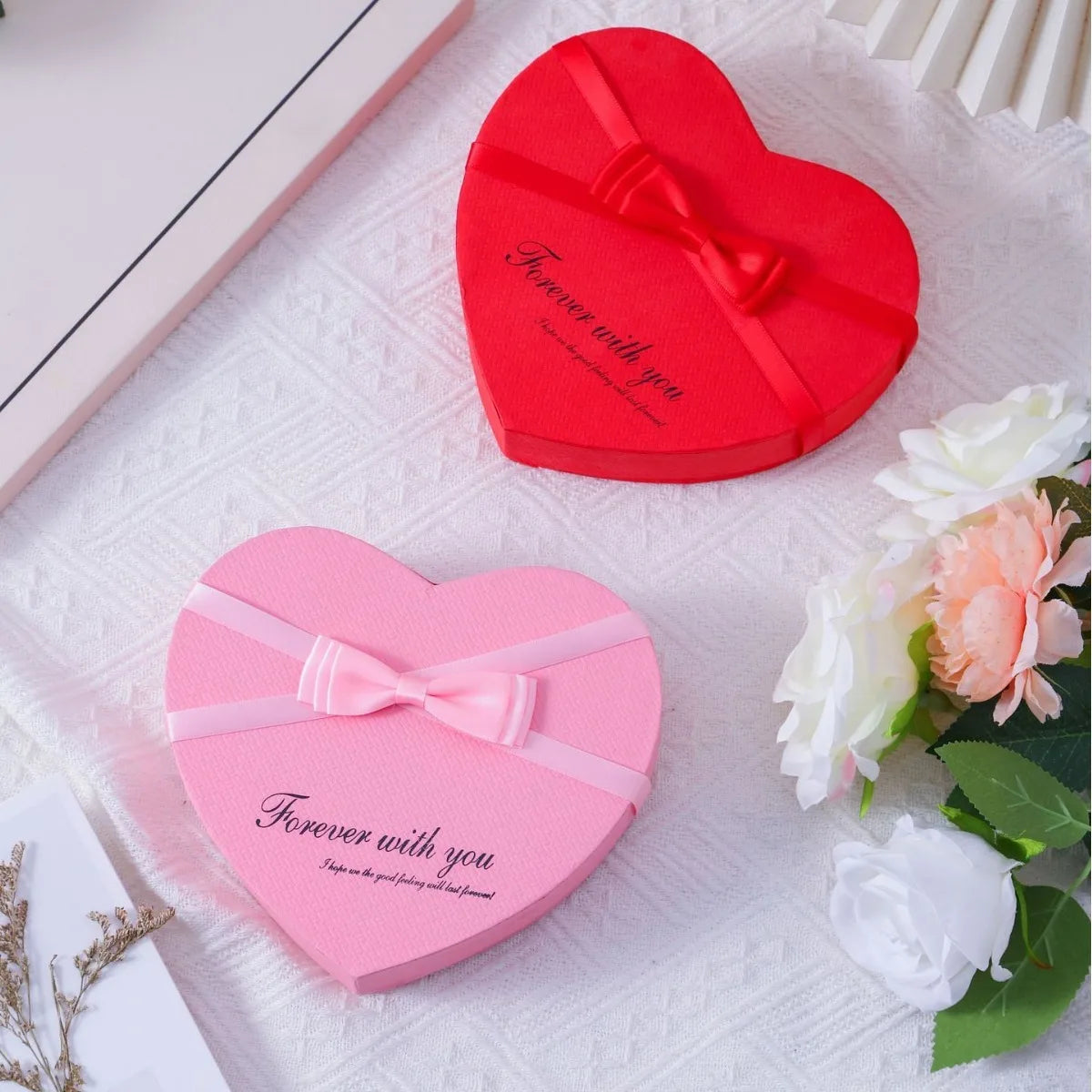 Red and pink heart-shaped gift boxes with ribbon and fresh roses displayed on a white surface