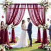 A couple stands in front of a draped floral archway with deep red curtains and greenery for an elegant event backdrop.