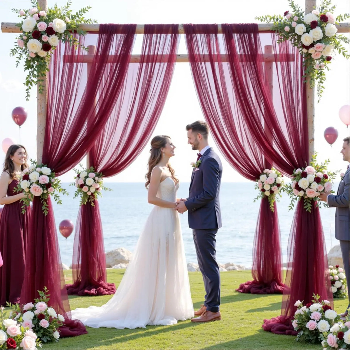 A couple stands in front of a draped floral archway with deep red curtains and greenery for an elegant event backdrop.