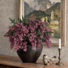 A bouquet of artificial nandina berry stems with dusty plum foliage in a dark vase on a wooden surface.