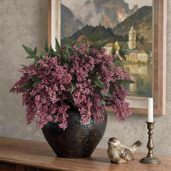 A bouquet of artificial nandina berry stems with dusty plum foliage in a dark vase on a wooden surface.