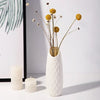 A white textured vase holds delicate dried flowers with thin stems and spherical yellow blooms.