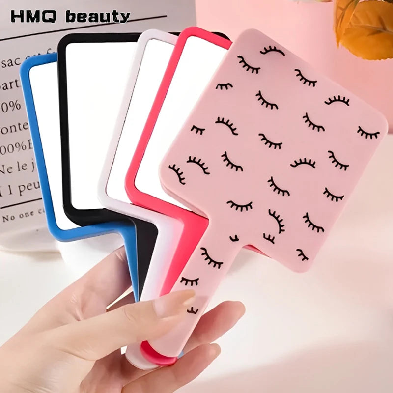 A hand holds a pink handheld vanity mirror with an ergonomic handle and several other colored mirrors.
