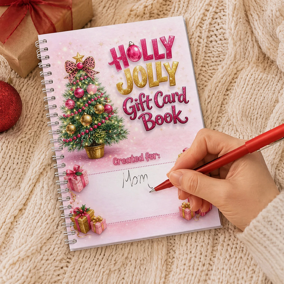 A hand writes in a pink and gold gift card book titled Holly Jolly Gift Card Book on a knitted background.