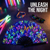 A purple and blue floral patterned folding rave hand fan is displayed on a dark surface with a neon light and sunglasses near