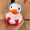 A white rubber duck bath toy with blue ears and red feet is held in a hand against a blurred background.