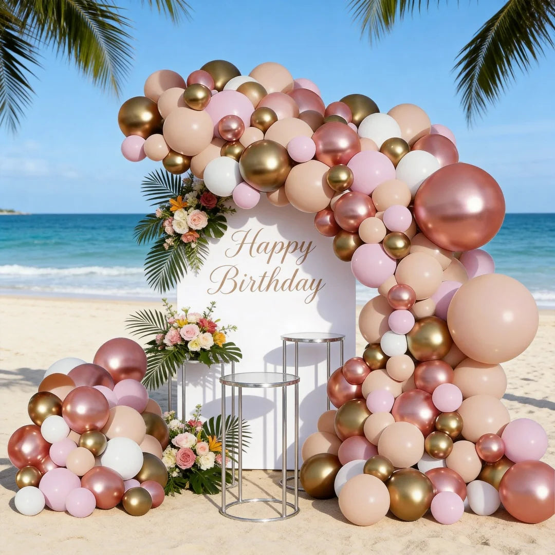 A large balloon garland featuring pink, rose gold, and cream balloons arranged in an arch shape on a beach.