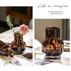 A dark amber leopard print glass vase with a pink rose and greenery on a white table.