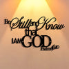 A black metal sign with elegant script text that reads Be Still and Know that I am God, set against a bright light background