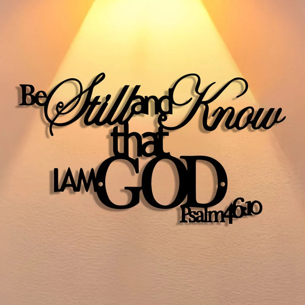A black metal sign with elegant script text that reads Be Still and Know that I am God, set against a bright light background