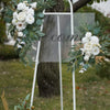 A white metal wedding arch decorated with white roses and green eucalyptus foliage in an outdoor setting.
