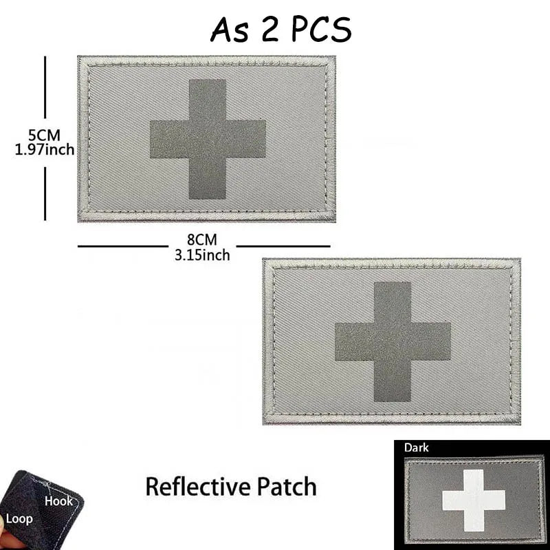 A rectangular reflective patch with a dark gray cross design and a loop for attachment.