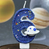 A blue glitter number six candle decorated with stars and a white planet design.