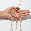A hand holds a gold moon-shaped crystal pendant with dangling rainbow prisms.