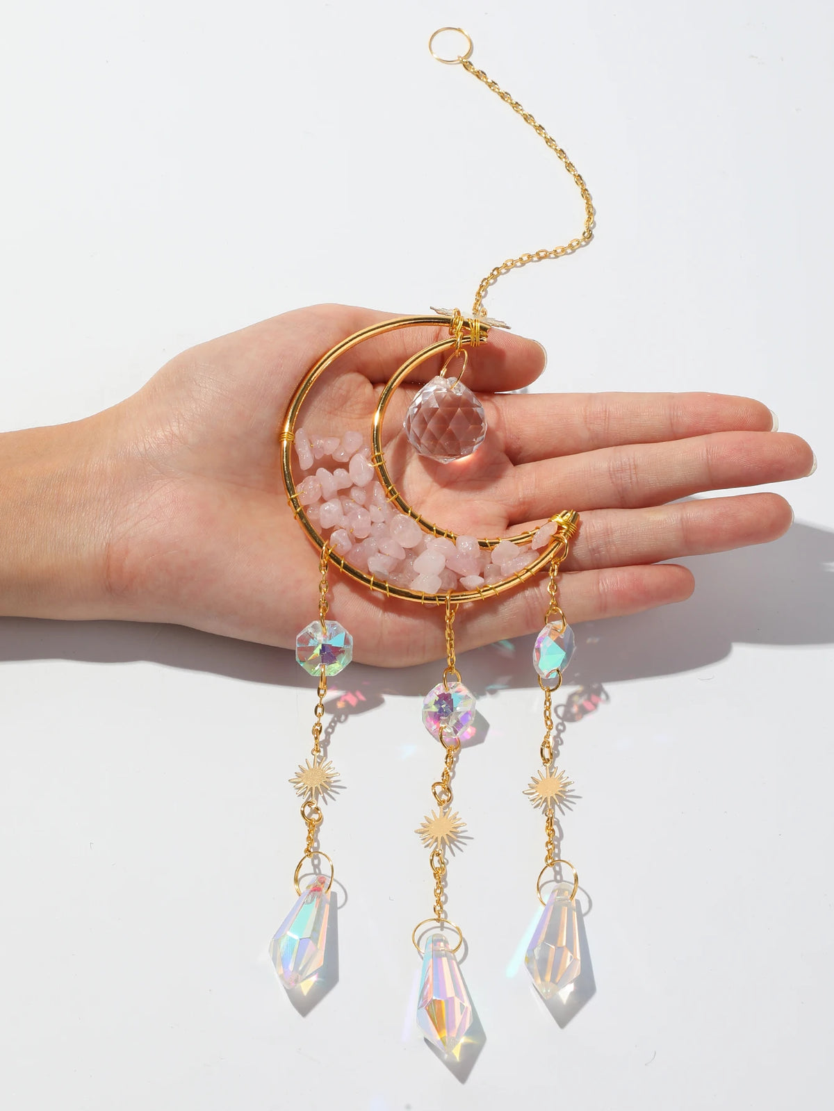 A hand holds a gold moon-shaped crystal pendant with dangling rainbow prisms.