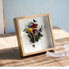 A wooden shadow box photo frame containing a bouquet of dark red and yellow flowers on a light wooden surface.