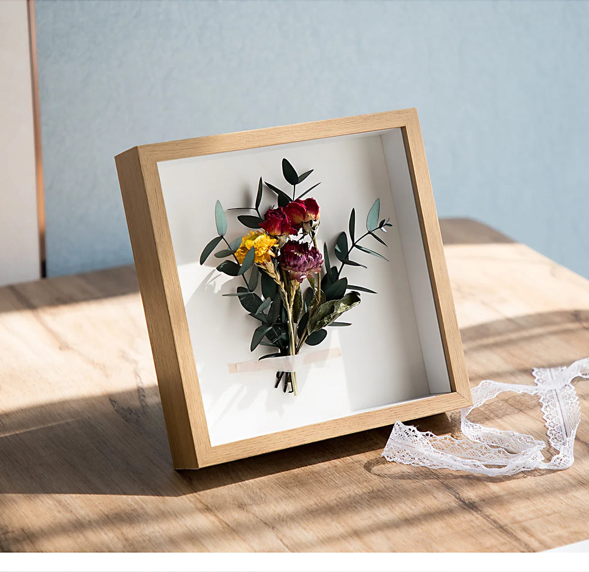 A wooden shadow box photo frame containing a bouquet of dark red and yellow flowers on a light wooden surface.