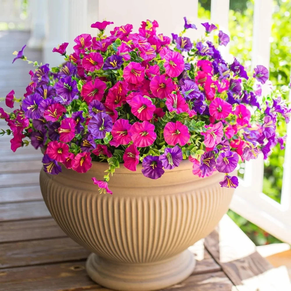 A vibrant bouquet of artificial flowers in shades of purple and pink arranged in a beige urn on a wooden porch.