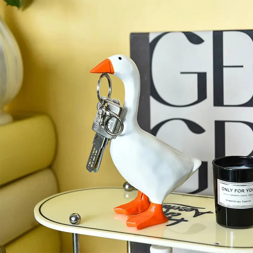 A white decorative resin figurine of a goose with an orange beak and feet, holding a set of keys.