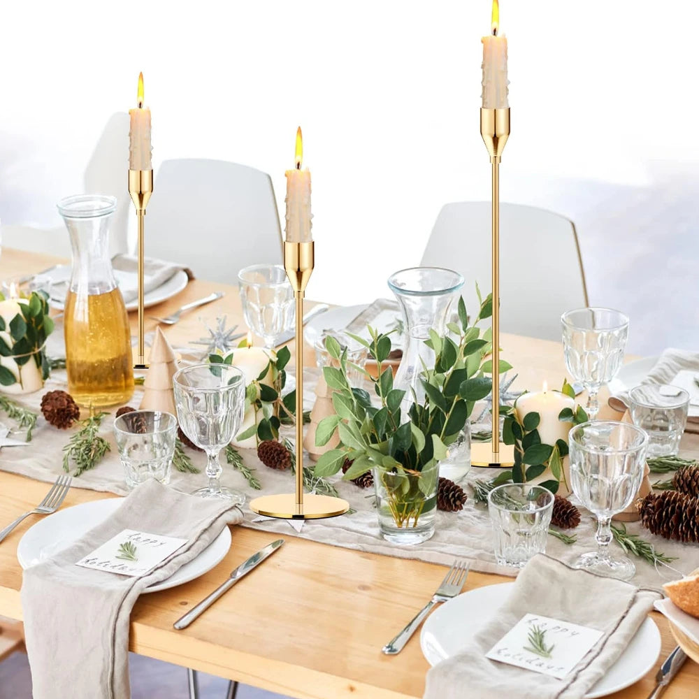 A set of three gold metal taper candle holders with white candles displayed on a wooden table setting.