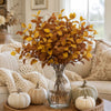 A bouquet of artificial fall eucalyptus stems in warm golden brown tones displayed in a clear glass vase on a woven mat.