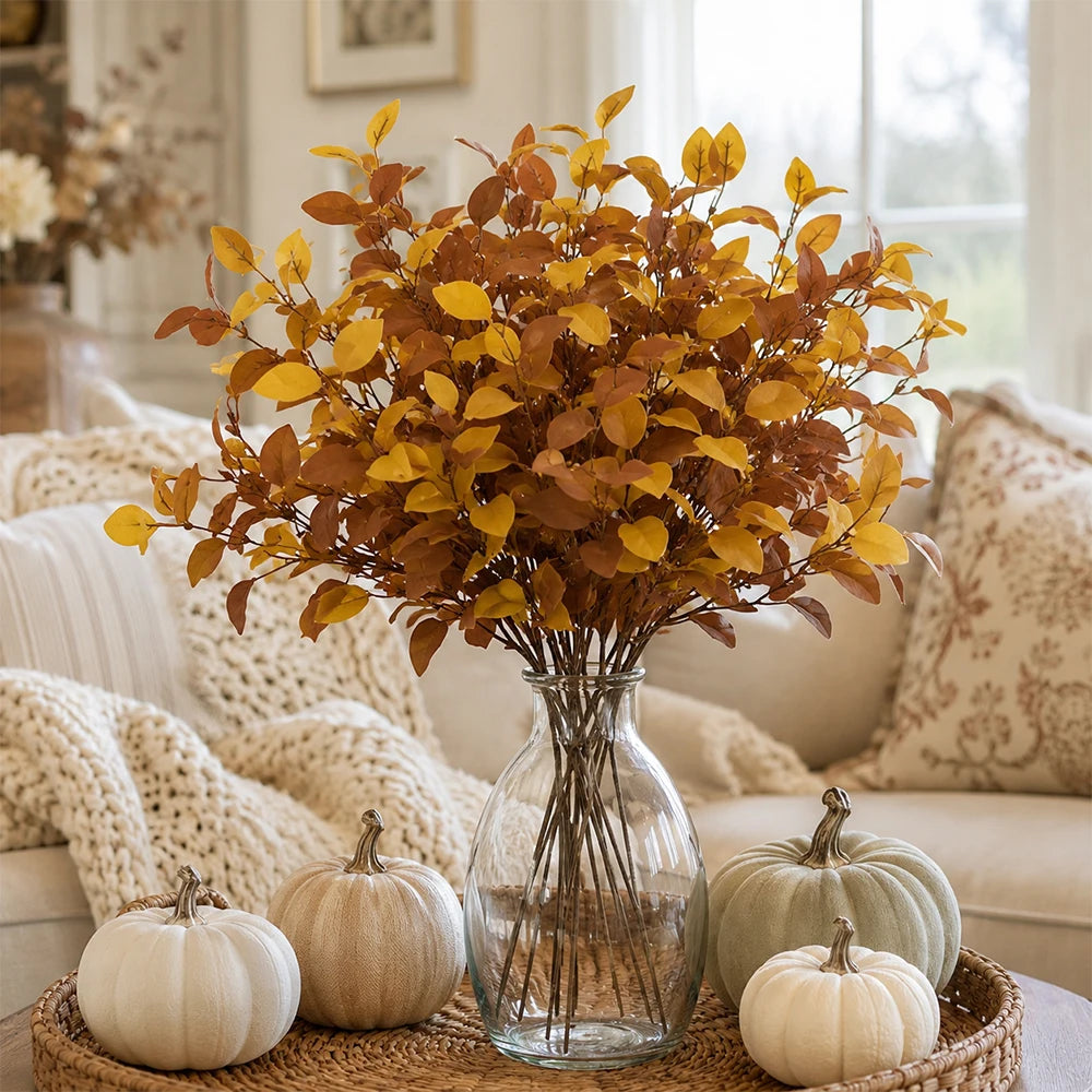 A bouquet of artificial fall eucalyptus stems in warm golden brown tones displayed in a clear glass vase on a woven mat.
