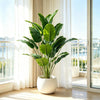 A vibrant green artificial Bird of Paradise tree in a white pot sits on a wooden floor next to a large window.