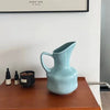 A light blue ceramic pitcher or vase sits on a wooden table next to small dark glass bottles.