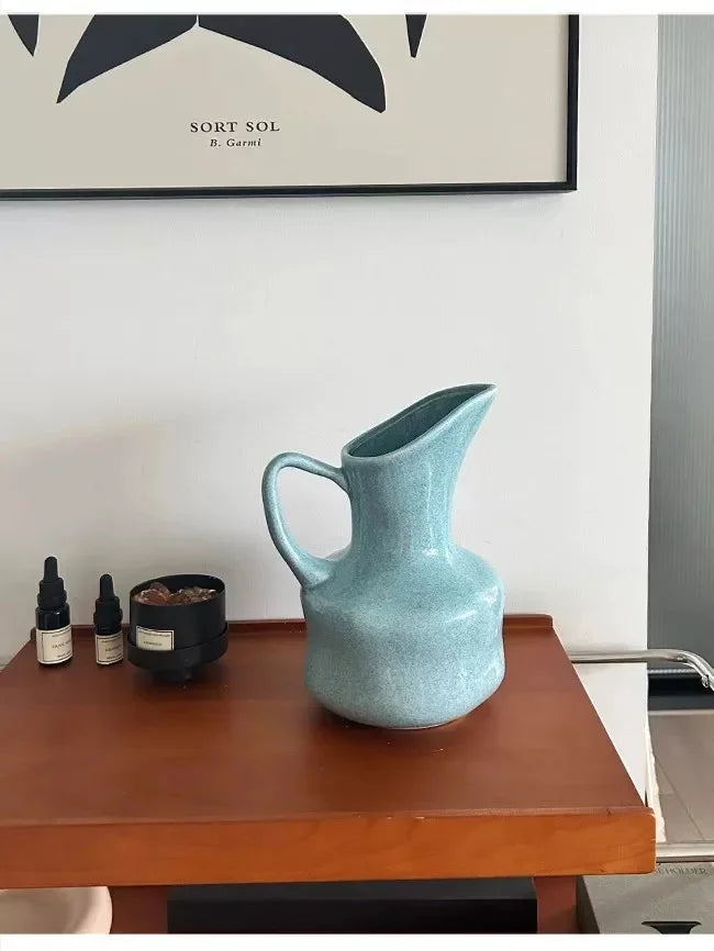 A light blue ceramic pitcher or vase sits on a wooden table next to small dark glass bottles.