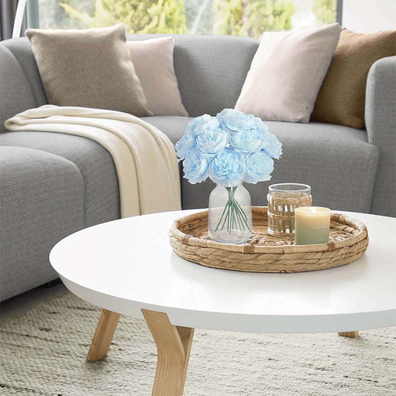 A glass vase filled with light blue artificial peony flowers sits on a woven tray on a white table.
