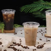 A glass of iced coffee or latte in a clear disposable plastic cup is placed on a white surface with coffee beans scattered ar