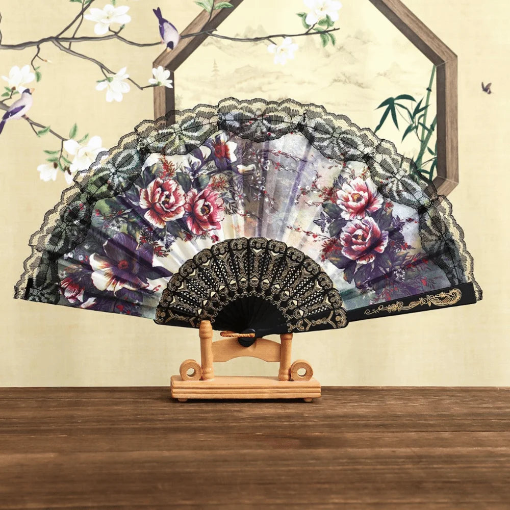 A Spanish style folding lace trim handheld fan with a dark floral pattern displayed against a light background.