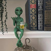 Resin green alien statue holding a glass of beer on a white shelf next to books