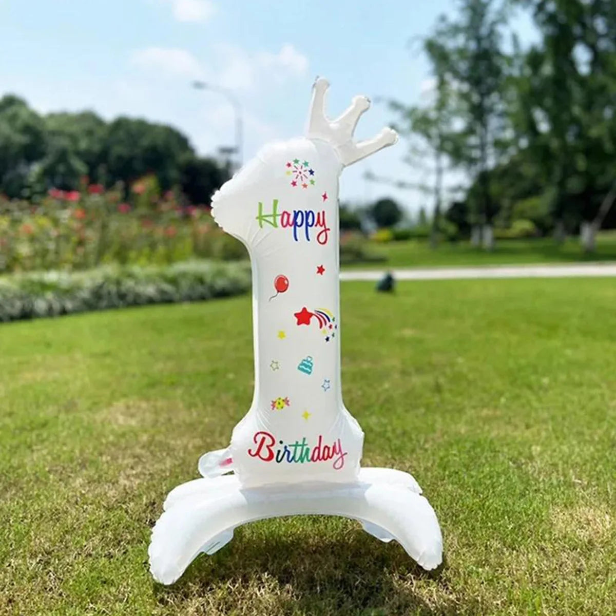 A white standing foil balloon shaped like a unicorn with a crown and birthday decorations on a green lawn.