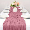 A pink boho style cheesecloth table runner draped over a white tablecloth with floral centerpiece and dinnerware.