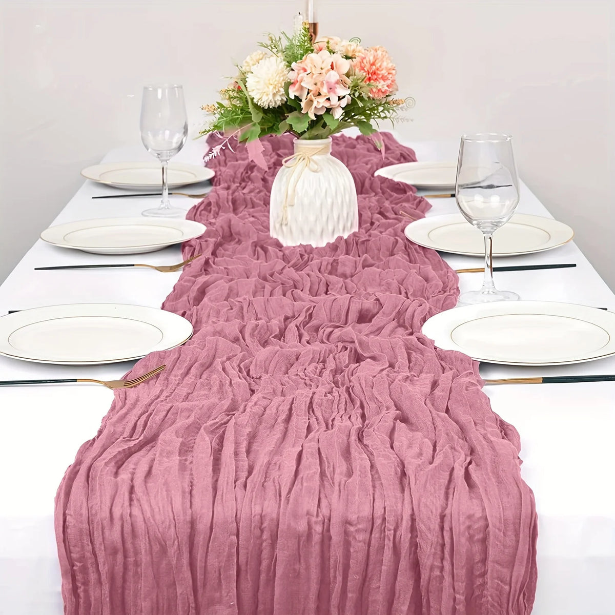 A pink boho style cheesecloth table runner draped over a white tablecloth with floral centerpiece and dinnerware.