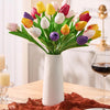 A bouquet of colorful artificial tulips in various shades of yellow, pink, red, and purple displayed in a white vase on a woo