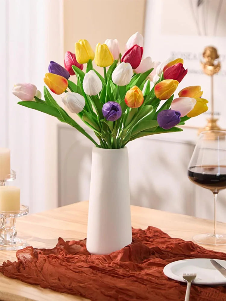 A bouquet of colorful artificial tulips in various shades of yellow, pink, red, and purple displayed in a white vase on a woo