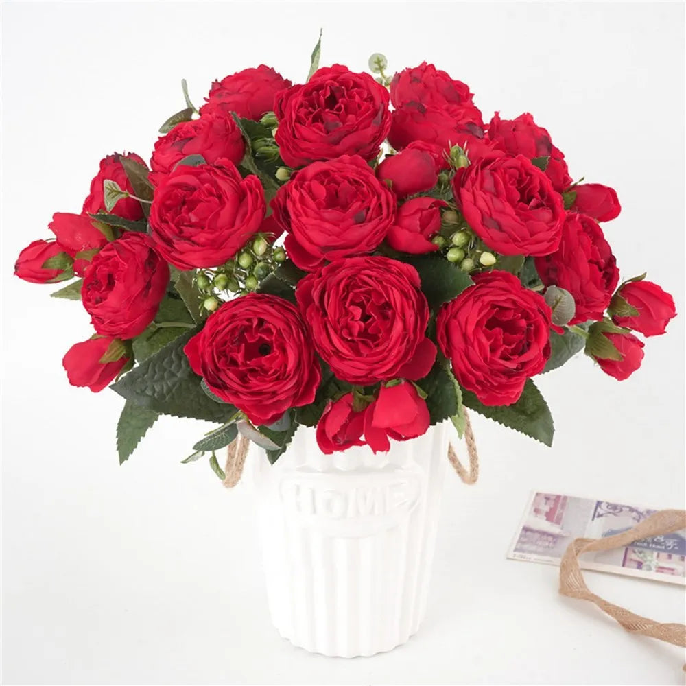 Ein Strauß künstlicher roter Rosen und Pfingstrosen in einer weißen zylindrischen Vase.