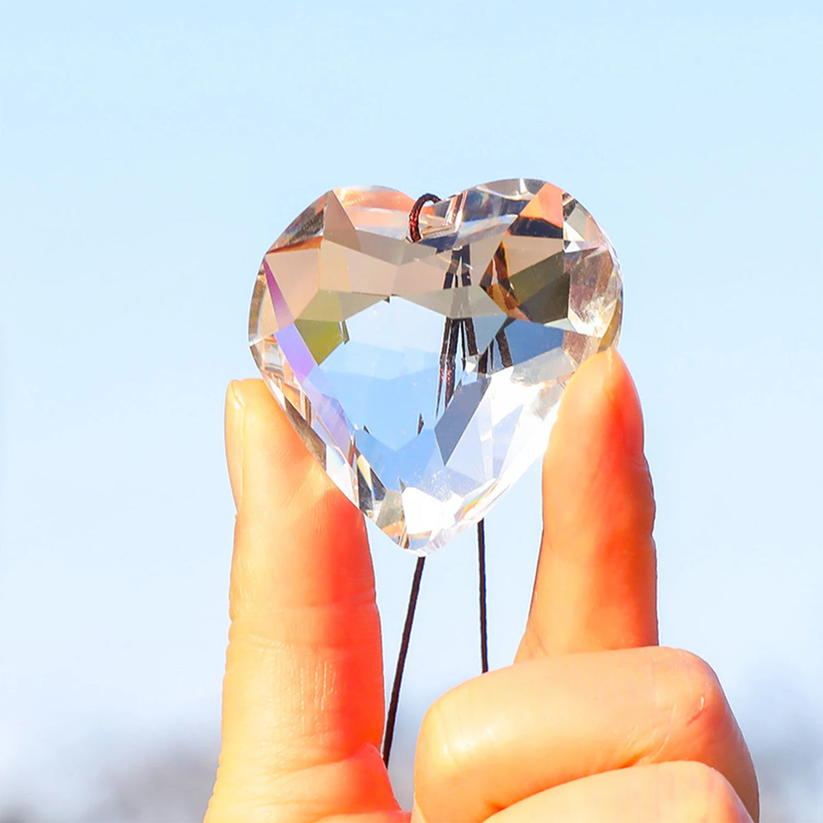 A hand holds a clear, faceted heart-shaped glass pendant catching the light against a pale blue sky.