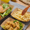 Disposable kraft paper food trays filled with fried chicken and potato wedges served on a gray cloth.