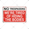 A vintage red metal warning sign with white text that reads NO TRESPASSING WE'RE TIRED OF HIDING THE BODIES