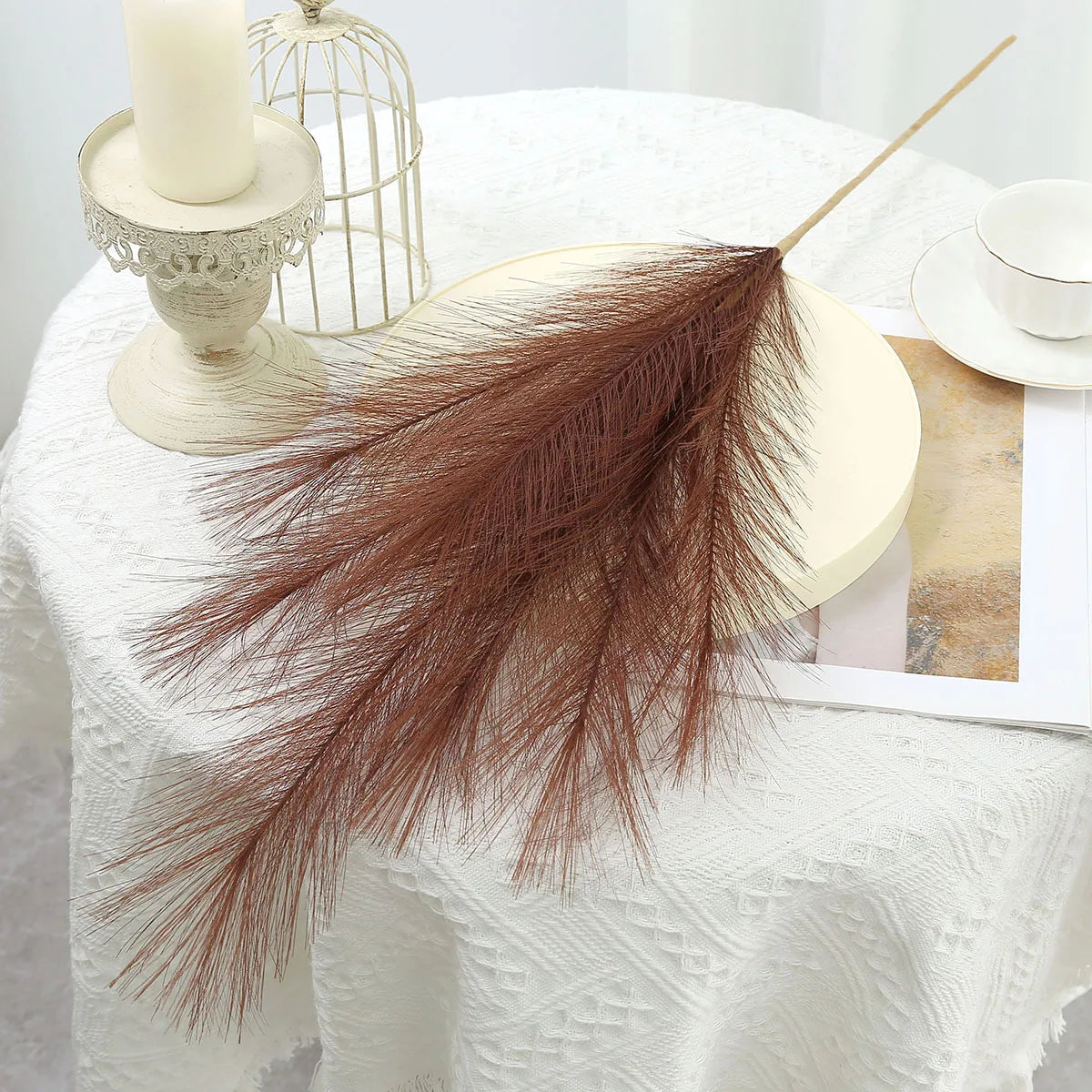 Dried brown pampas grass stalks arranged on a white linen tablecloth with a candle and wire cage
