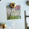A collection of gold star-shaped fridge magnets and a sign reading ICE CREAM on a white surface.
