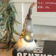 A knitted gray and white LED gnome ornament with a glowing face sits next to a small green tree.