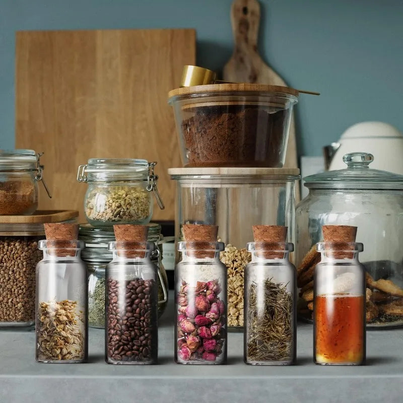 A collection of decorative glass vials filled with various dried herbs, spices, and grains arranged on a gray surface.