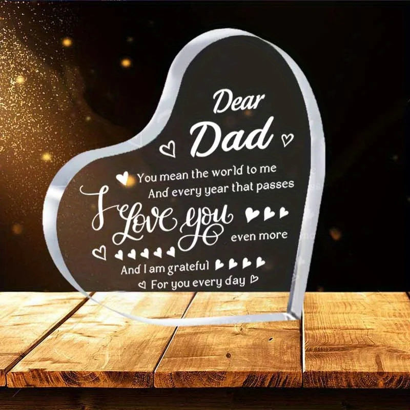 A clear acrylic heart plaque engraved with 'Dear Dad I love you even more' resting on a wooden surface.